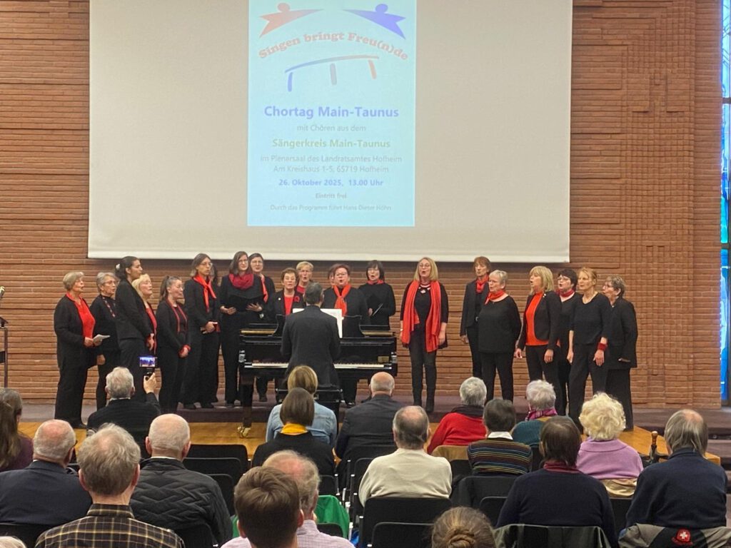 Der Frauenchor Lorsbach singt beim Chortag Main-Taunus im Plenarsaal des Landratsamtes Hofheim. Auf der Bühne steht der Chor in schwarzer Kleidung mit roten Akzenten, im Hintergrund ist ein Plakat mit dem Motto „Singen bringt Freude“ zu sehen. Vor der Bühne sitzen zahlreiche Zuhörer.