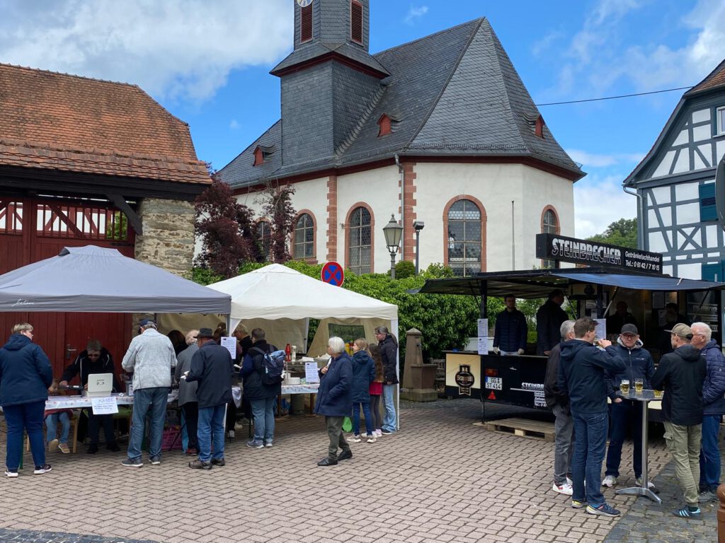 Besucherinnen und Besucher des Sängerfrühschoppens am Vatertag auf dem Zimmerplatz in Lorsbach. Vor der evangelischen Kirche werden Speisen und Getränke angeboten, darunter Grillgut und frisch gezapftes Bier.