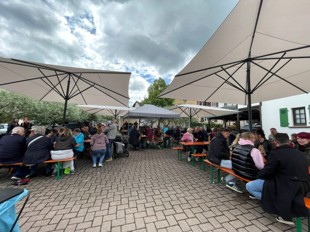 Besucherinnen und Besucher sitzen beim Sängerfrühschoppen am Vatertag auf dem Zimmerplatz in Lorsbach gemeinsam an Bierbänken unter großen Sonnenschirmen. Die Gäste verbringen den Nachmittag bei Essen, Getränken und Gesprächen im Freien.